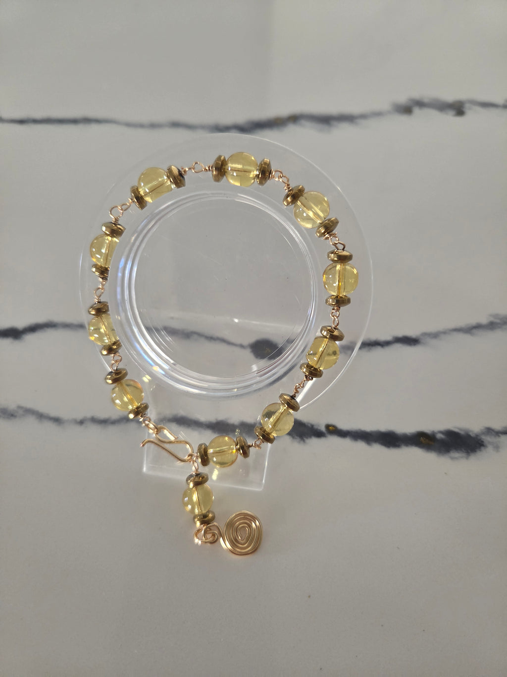 A beaded stretch bracelet with faceted yellow beads and rose gold-toned metal spacers and charm, displayed on a clear acrylic stand over a white marble-patterned surface.