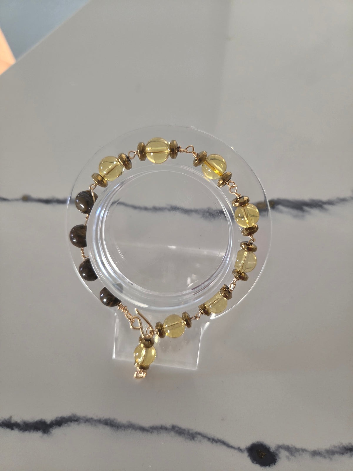 A beaded bracelet displayed on a clear acrylic stand, featuring yellow translucent beads, dark brown beads, and gold-toned metal links and charms, set against a light gray marble-patterned background.