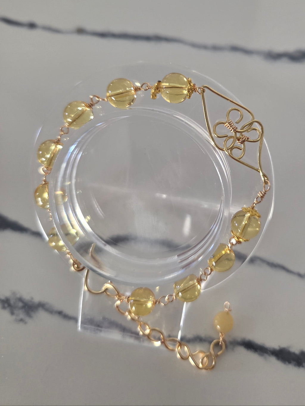 A gold-tone bracelet with faceted yellow beads and a geometric flower motif, displayed on a clear acrylic stand on a light marble surface.