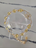 A gold-tone bracelet with faceted yellow beads and a geometric flower motif, displayed on a clear acrylic stand on a light marble surface.