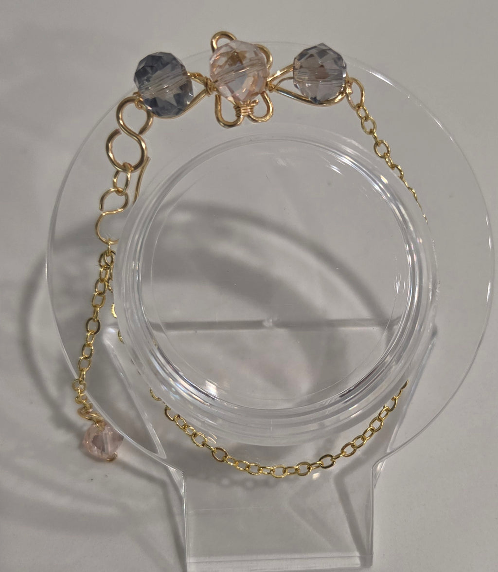 A delicate gold chain bracelet with three faceted beads in light blue, pale pink, and pale lavender, displayed on a clear acrylic jewelry stand.
