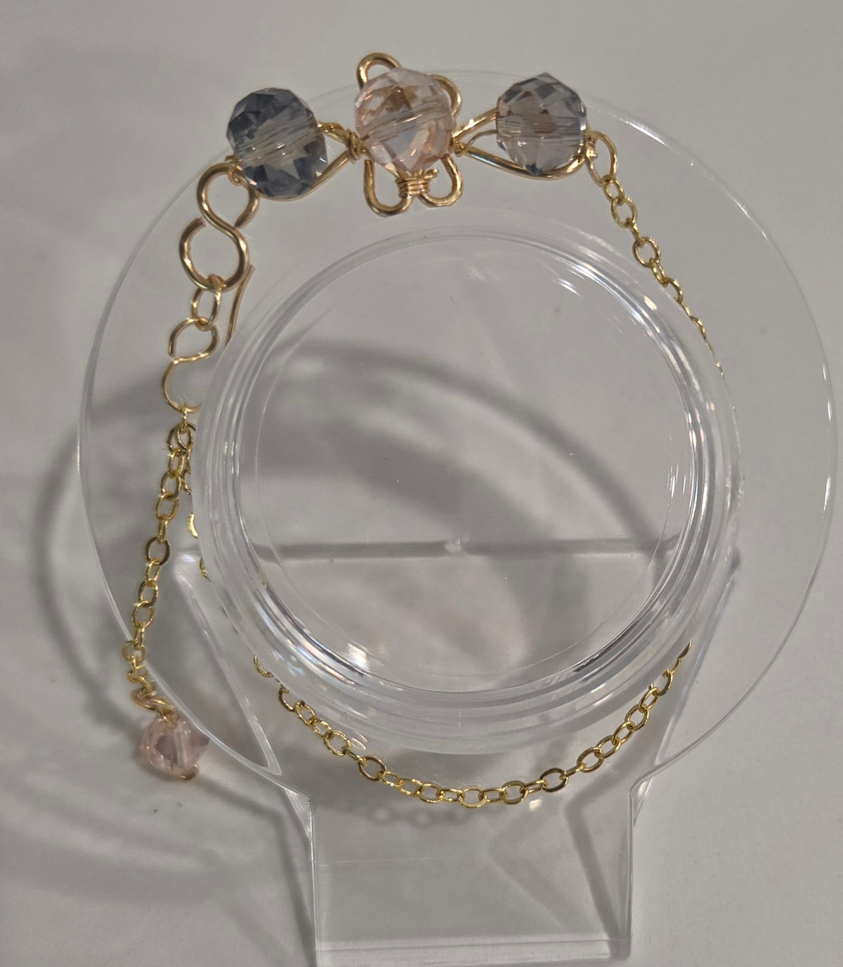 A delicate gold chain bracelet with three faceted beads in light blue, pale pink, and pale lavender, displayed on a clear acrylic jewelry stand.
