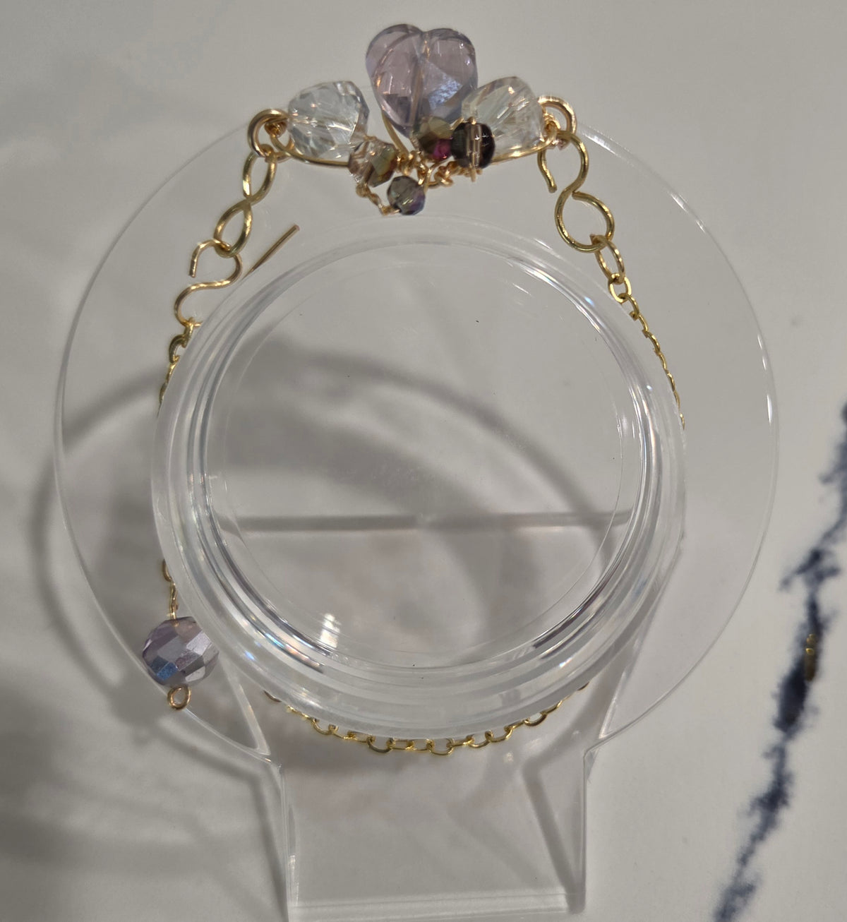 A delicate gold chain bracelet with faceted crystal and lavender beads, displayed on a clear acrylic stand against a light gray background.