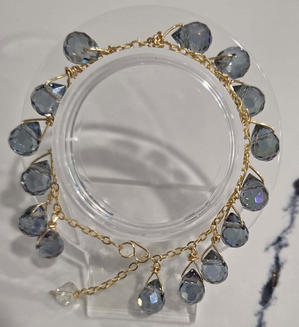 A gold chain bracelet with dangling faceted gray-blue teardrop crystals, displayed on a clear acrylic stand against a light neutral background.