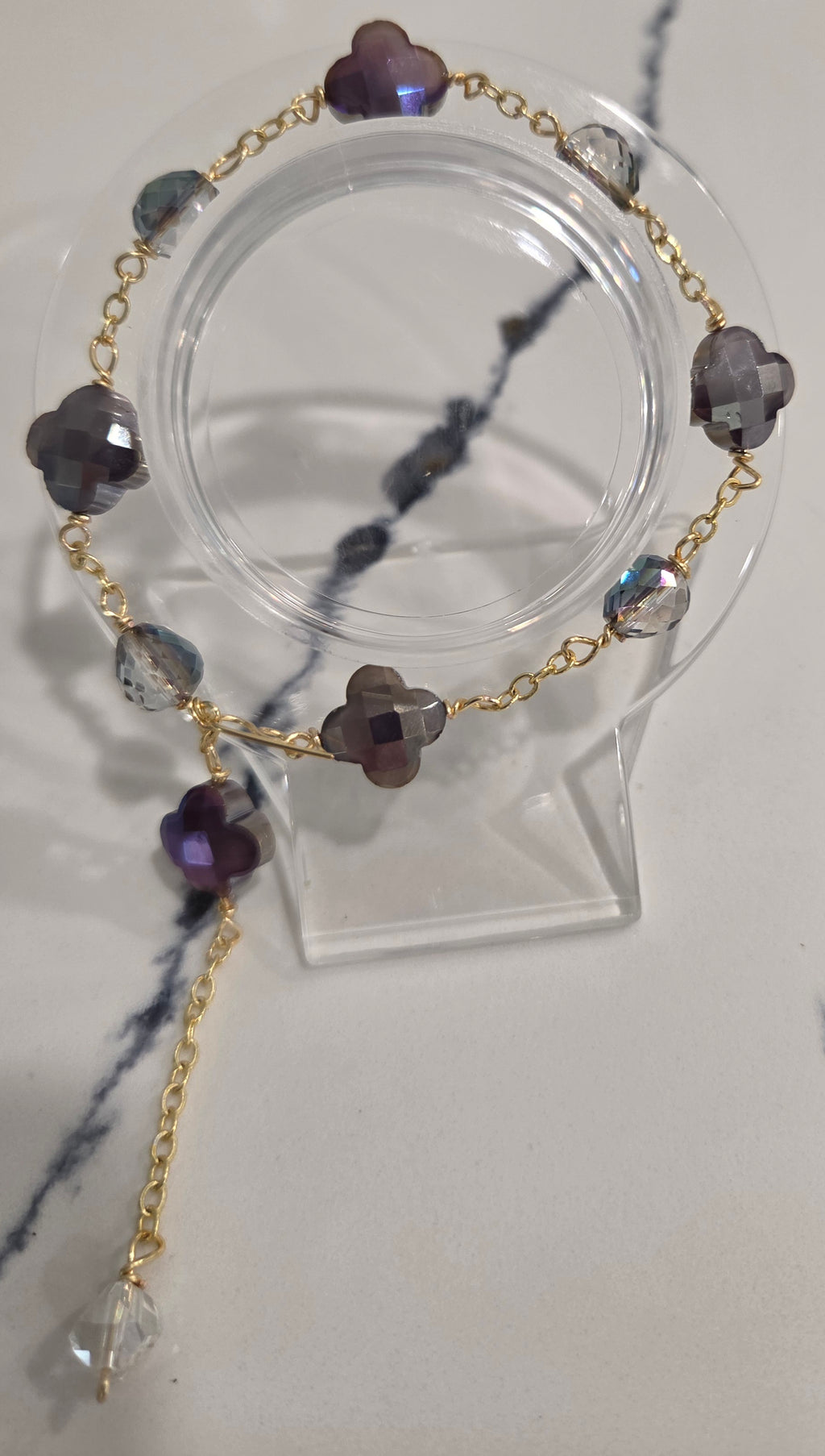 A gold chain bracelet with faceted purple and gray beads, displayed on a clear acrylic stand against a light marble-patterned background.