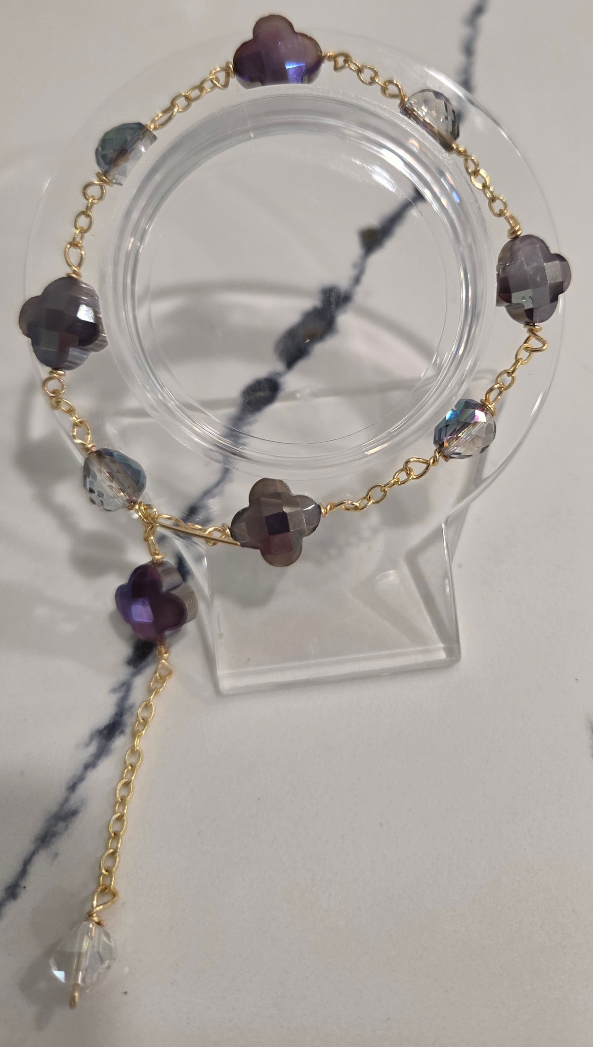 A gold chain bracelet with faceted purple and gray beads, displayed on a clear acrylic stand against a light marble-patterned background.