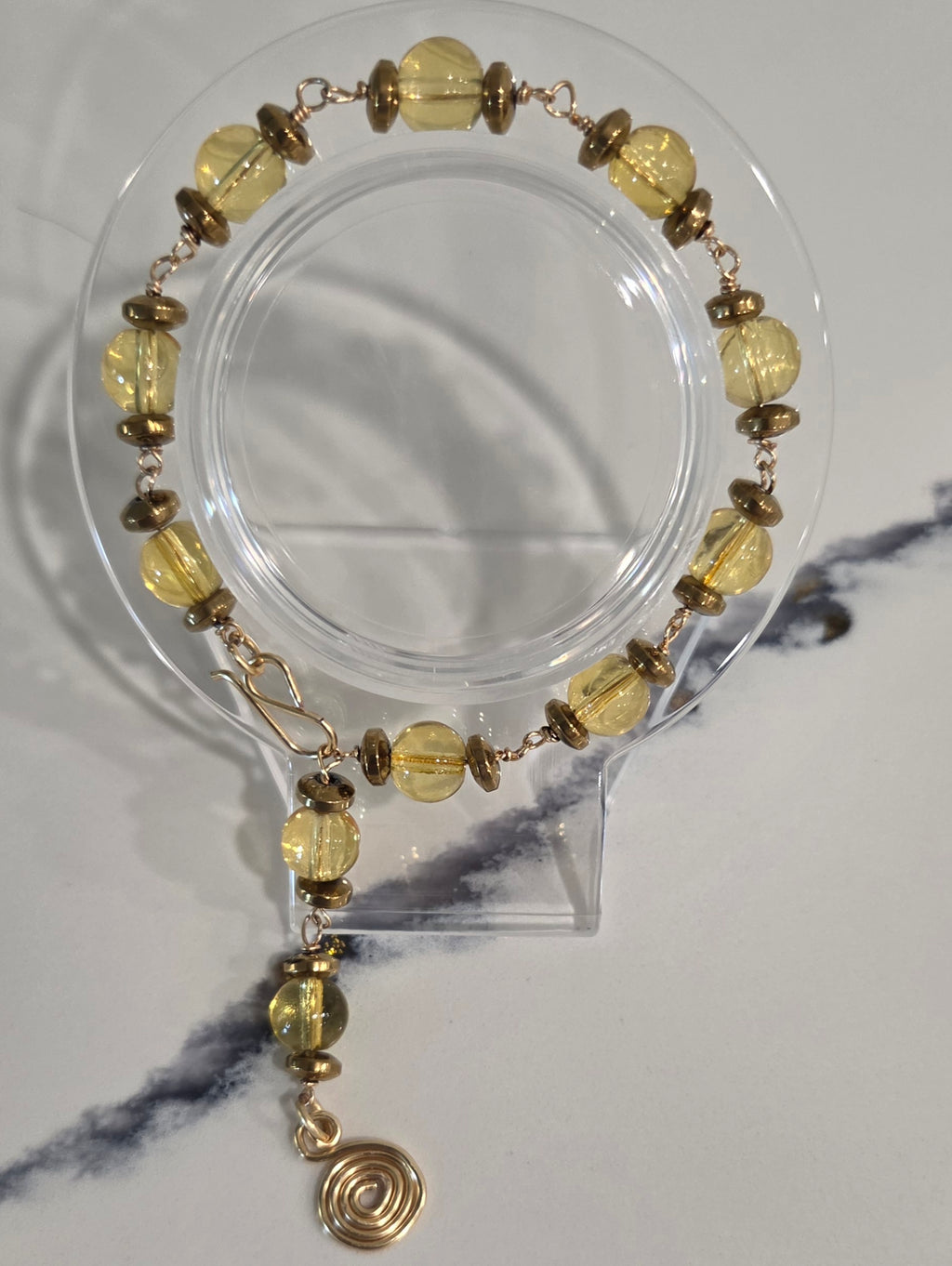A beaded bracelet with translucent yellow-green faceted beads and small metallic gold spacer beads, displayed on a clear acrylic stand against a light gray marble-patterned background.