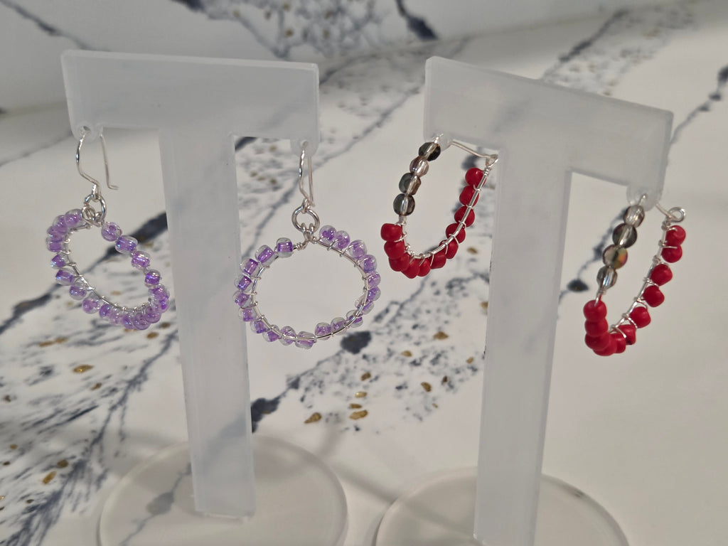 Three pairs of handmade beaded hoop earrings displayed on clear acrylic stands. The left pair consists of purple beads, the middle pair of red and black beads, and the right pair of red beads with silver-toned metal findings.