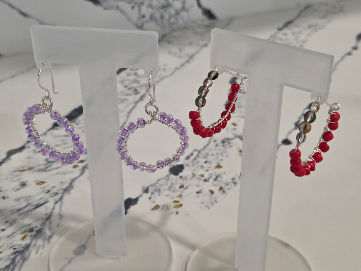 Three pairs of handmade beaded hoop earrings displayed on clear acrylic stands. The left pair consists of purple beads, the middle pair of red and black beads, and the right pair of red beads with silver-toned metal findings.